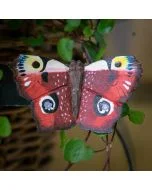 Houten DecoButterfly Dagpauwoog van Wildlife Garden met roodbruine vleugels en opvallende blauwgele oogvlekken, bevestigd tussen groene plantenbladeren.