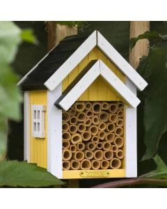 Biholk Geel bijenhotel van Wildlife Garden tussen groene bladeren in de tuin, met gele houten gevel, witte dakranden, zwart dak en holle bamboebuisjes.