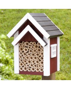 Biholk Rood bijenhotel van Wildlife Garden op een houten paal in een groene tuin, met rode gevel, witte dakranden, donker dak en holle nestbuisjes.