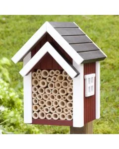 Biholk Rood bijenhotel van Wildlife Garden op een houten paal in een groene tuin, met rode gevel, witte dakranden, donker dak en holle nestbuisjes.