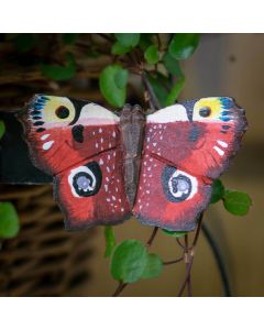 Houten DecoButterfly Dagpauwoog van Wildlife Garden met roodbruine vleugels en opvallende blauwgele oogvlekken, bevestigd tussen groene plantenbladeren.