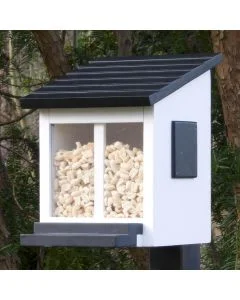 Eekhoorn voederhuisje Ekorrmatare Wit van Wildlife Garden op een paal tussen bomen, met zwart schuin dak en lichte voerstukjes achter de transparante vensters.