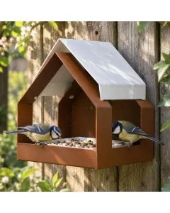 The Barn wandvoederhuis bruin van metaal met wit zadeldak, gemonteerd aan een houten schutting, waar twee koolmezen vogelvoer eten.