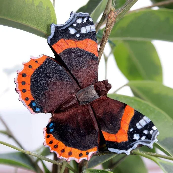 DecoButterfly Atalanta van Wildlife Garden als houten vlinderdecoratie tussen groene bladeren, met donkerbruine vleugels, oranjerode banden en witte vlekken.
