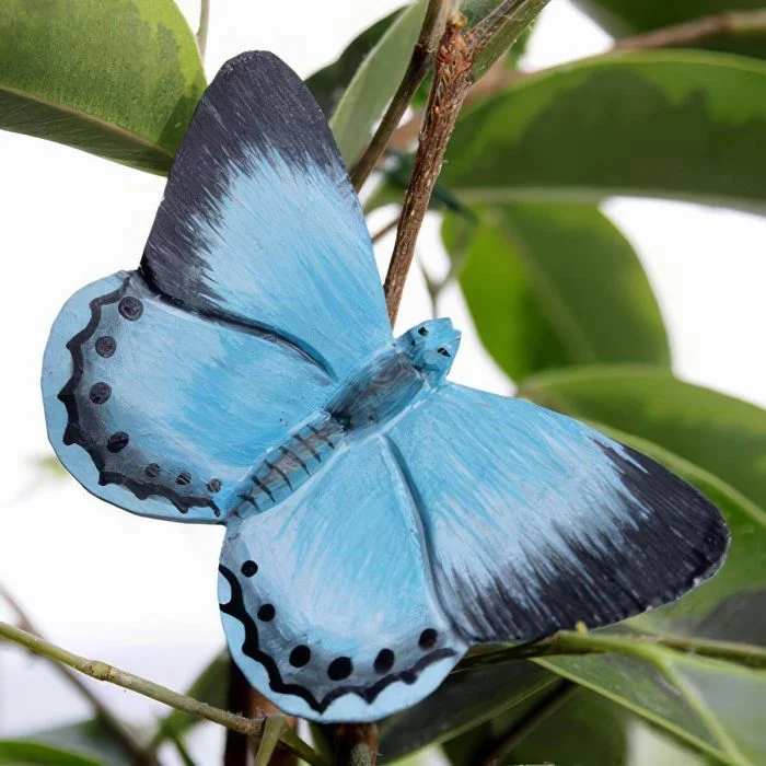 DecoButterfly Boomblauwtje van Wildlife Garden als houten vlinderdecoratie tussen groene bladeren, met lichtblauwe vleugels, zwarte vleugelpunten en donkere stippen.
