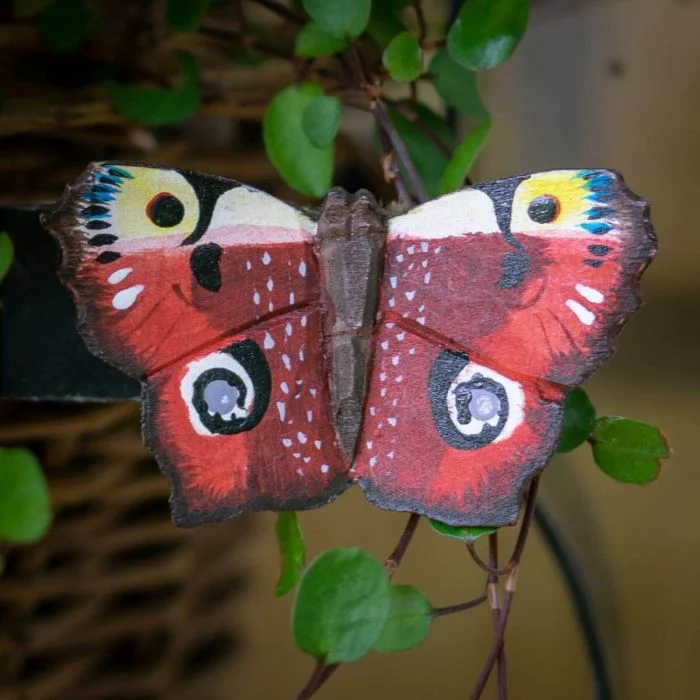Houten DecoButterfly Dagpauwoog van Wildlife Garden met roodbruine vleugels en opvallende blauwgele oogvlekken, bevestigd tussen groene plantenbladeren.