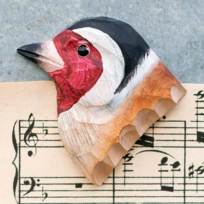 Houten koelkastmagneet Putter van Wildlife Garden met rode gezichtstekening, zwart-witte kop en bruine vleugel, gefotografeerd op bladmuziek.